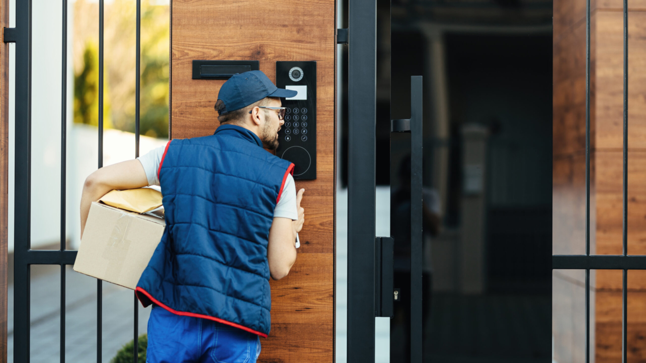 Back view of a deliverer ringing on intercom at gate of a customer's house while delivering packages.